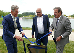 Tafel, die an die Widmung von Herrn Herrenknecht erinnert. Steht im Seepark und ist mit einem blauen Band umwickelt. 
An dem Band ziehen gemeinsam OB Dr. Müller und Altkanzler Schröder. Herrenknecht schaut in der Mitte stehend auf die Tafel. 