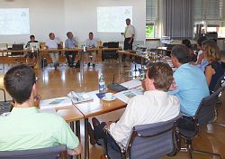 Bürgermeister Tilman Petters steht vor einer Leinwand im Gemeinderatssaal und begrüßt die Teilnehmer, die ihm halbkreisförmig zugewandt an Tischen sitzen.