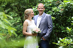 Ein Hochzeitspaar steht im Grünen. Sie trägt ein weißes Spitzenkleid und lächelt ihn von unten an. Er hat lässig die Hand in der Hüfte gestemmt, trägt einen blauen legeren Anzug mit weißem Hemd. 