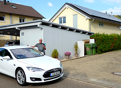 Ein weißer Tesla steht an einer Straße am Hang. Dahinter Alexander Schanze mit einem Kabel für das E-Auto. Im Hintergrund steht ein Haus mit Photovoltaikanlage auf dem Dach. 
