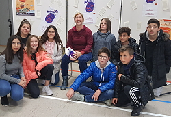 Die Autorin Stefanie Höfler sitzt inmitten von Jugendlichen und hält ihr Buch in die Kamera. Im Hintergrund eine Stellwand mit herausgetrennten Buchseiten und einem Plakat mit dem Cover des Buches "Tanz der Tiefseequalle". 