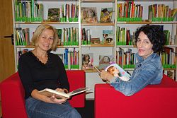 Das Foto zeigt eine gemütliche Sitzecke in der Mediathek, im Hintergrund ein großes Bücherregal. Links in einem knallroten Sessel Birgit König, rechts Diana Dold. Beide haben ein aufgeschlagenes Buch in den Händen.