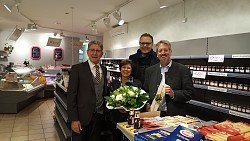 Die Lahrer Verwaltungsspitze bedankt sich bei Otlilia-Brüderle, in einer weinroten Schürze, mit einem Blumenstrauß. Sie stehen vor der Kühlvitrine des Geschäfts. 