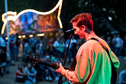 Vincent Alexander steht erhöht auf einer Bühne und spielt auf seiner Gitarre. Er ist in rotes Licht getaucht, das Publikum unter ihm leuchtet in blauem Licht.