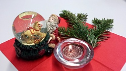 Auf einer roten Papierserviette brennt ein Teelicht mit Glitzerstaub. Dahinter ein Tannenzweig. Links eine Schneekugel in Form eines Weihnachtsmannes, der seinen Geschenkesack öffnet und Geschnke zu sehen sind.