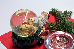 Auf einer roten Papierserviette brennt ein Teelicht mit Glitzerstaub. Dahinter ein Tannenzweig. Links eine Schneekugel in Form eines Weihnachtsmannes, der seinen Geschenkesack öffnet und Geschnke zu sehen sind.