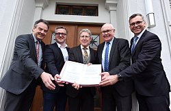Vor dem Rathaus stehen fünf Herren und halten gemeinsam den Vertrag in den Händen, den sie lächelnd in die Kamera halten. 