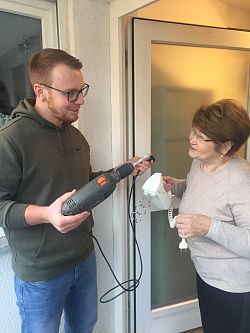 Ein junger Mann mit einer Bohrmaschine in der Hand steht vor der offenen Türe einer Frau. Diese hält einen Mixer in der Hand.
