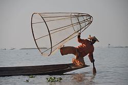 Auf einem See ist ein Fischerboot, darauf steht ein einheimischer Fischer in schon fast akrobatischer Haltung am Ende des Bootes und wirft einen Korb aus. 