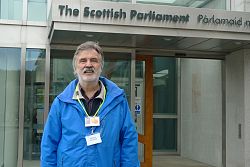 Mann mit grau-melierten Haaren, grauem Schnauzbart und grauem 7-Tage-Vollbart, trägt blaue Jacke und steht vor einem Gebäude mit der Aufschrift "The Scottish Parliament".