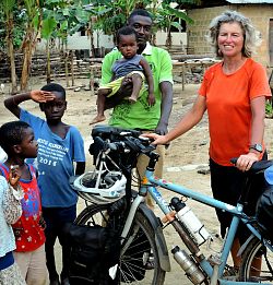 Auf dem Bild steht Dorothee Fleck rechts mit ihrem Fahrrad, links von ihr ein Mann aus Ghana mit einem kleinen Jungen auf dem Arm, neben ihm zwei kleine Jungs