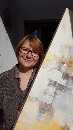 Das Bild zeigt die Künstlerin, die einen braunen Pulluvover trägt, und eines in sanften Erdtönen gestaltetes Bild schräg in Händen hält. 