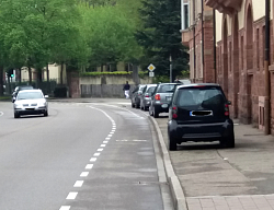 Auf dem Bild sind vier graue und schwarze Autos zu sehen, die rechts auf dem Gehweg neben einem alten Gebäude parken. Auf der linken Spur fährt ein silbernes Auto. Im Hintergrund sieht man mehrere große Bäume.