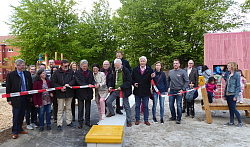 Über den Spielplatz ist ein rot-weißes Flatterband gespannt, das von Vertretern des Gemeinderats und der Stadtverwaltung durchschnitten wurde. In der Mitte steht lächelnd OB Dr. Müller mit der Schere. 