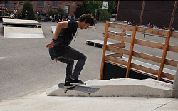 Zu sehen ist ein junger Mann, der mit einem Skateboard eine Rampe hoch springt. Er trägt schwarze Kleidung und Kopfhörer. Im Hintergrund sind weitere Rampen und Zuschauer zu sehen.