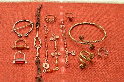 Zu sehen sind viele filigrane Schmuckstücke aus Perlen und Drähten. Sie sind in warmen, goldenen Farbtönen gehalten und werden auf einem roten Tuch präsentiert.