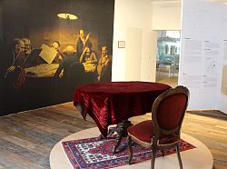 Das Bild zeigt einen Holztisch mit einer großen roten Samttischdecke und einen Stuhl aus Holz, ebenfalls bezogen mit rotem Samt auf einem bunten Teppich plaziert in einem extra Raum im Stadtmuseum. 