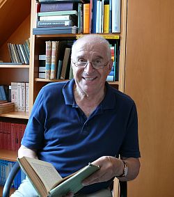Zu sehen ist Prof. Dr. Heinz Kneile in seinem Büro. Er trägt ein dunkelblaues Polo-Shirt und eine Brille. In der Hand hält er ein Buch, im Hintergrund ist ein voll bestücktes Bücherregal zu sehen.