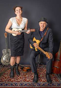 Das ist ein Porträtfoto von Kabarettistin Nessi Tausendschön. Sie trägt ein weiß-schwarzes Kleid, zwei Zöpfe und in der Hand hat sie eine Ukulele. Neben ihr sitzt ihr Showpartner mit Gitarre auf dem Schoß. Die Wand hinter ihnen ist schwarz, unter ihnen liegt ein roter Perserteppich.