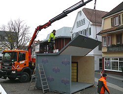 Zu sehen sind zwei BGL Mitarbeiter beim Abbau der Chrysanthema Häuschen auf dem Urteilsplatz. Ein Mitarbeiter steht auf dem Häuschen, während der vordere Bereich des Daches vom Kranausleger eine LKWs angehoben wird.