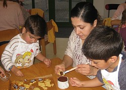 Eine Mutter sitzt mit zwei Kindern an einem Tisch und bestreicht mit einem Pinzel und Schokolade Weihnachtsgebäck. 