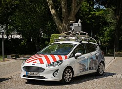 Zu sehen ist ein Auto der Firma cyclomedia, das zur Straßenbefahrung genutzt wird. Für die Panaoramaaufnahmen und Scans hat das Fahrzeug eine Kameraausrüstung auf dem Dach. Das Auto ist ein Kleinwagen in der Farbe weiß. Auf der Kühlerhaube sind breite rote Warnstreißen, wie man sie von Baustellenabsperrungen kennt, aufgebracht. Seitlich am Auto ist ein Foto einer Stadtansicht aufgeklebt.