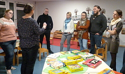 Eine Erzieherin steht an einem Tisch, auf dem mehrere Spiele aufgebaut sind. Darum gruppieren sich Eltern und hören aufmerksam der Erzieherin zu. 