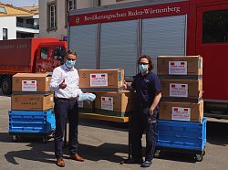 Oberbürgermeister Markus Ibert und Feuerwehrkommandant Thomas Happersberger stehen vor einem Tisch und zwei Rollboxen, auf denen Kartons mit Schutzmasken aus China gestapelt sind. Dahinter sind Auto der Feuerwehr Lahr zu sehen. Beide Männer tragen eine Maske aus der Lieferung über Nase und Mund. Sie strecken einen Daumen nach oben als Zeichen für &#34;Gefällt mir!&#34;
