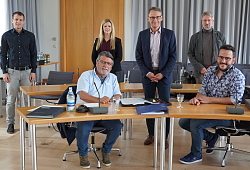 Im Gemeinderatssaal sitzen im Vordergrund Lothar und Marc Scheer an den Tischen, vor sich verschiedene Ordner. Im Hintergrund haben sich von links nach rechts Stefan Martinelli, Silke Kabisch, OB Ibert und Bürgermeister Petters im Abstand verteilt. Alle lächeln in die Kamera. 
