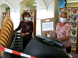 Karin Schmidt und Birgit König von der Mediathek Lahr stehen vor dem Zeitschriftenregal. Schmidt lehnt sich an einen Stapel Stühle und Birgit König steht hinter gestapelten Sesseln neben einem Schild auf dem steht, wir freuen uns wenn Sie wieder hier sitzen können. 