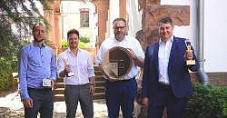 Von links stehen Jacob Crone (Stadt Lahr), Pierre Grieger (Vollblutwerber), Erster Bürgermeister Guido Schöneboom und  Malte C. Bayer (Vollblutwerber. Sie halten jeweils Pokale und Schöneboom einen überdimensionalen Stadtgulden in den Händen. Sie stehen im Freien, im Hintergrund eine alte Villa, der Firmensitz der Vollblutwerber