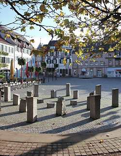 Blick auf den Marktplatz, im Vordergrund stehen die Skulpturen. Graue, freistehende und unterschiedlich hohe Granitstelen sind kreisförmig aufgestellt. 