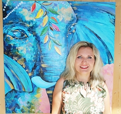 Die blonde Künstlerin steht lächelnd in einem Kleid mit Dschungeldruck vor ihrem Bild &#34;Das Elefantenfesival in Indien&#34;. Man sieht einen übergroßen Elefantenkopf in blau gehalten, der mit einem bunten Zweig von Blättern geschmückt ist. 