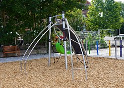 Ein Klettergerät auf dem Pausenhof Hort Luisenschule steht auf einem Platz mit Holzschnitzeln als Fallschutz. Im Hintergrund ist ein Metallzaun mit bunten Zaunguckern aus Holz zu sehen. Das Gerät hat eine kleine Kletterwand, einen grünen Schaukelsitz, ein Netz, Stangen und eine geschwungene Leiter.