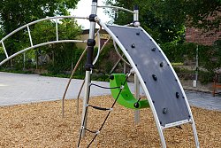 Ein Klettergerät auf dem Pausenhof Hort Luisenschule steht auf einem Platz mit Holzschnitzeln. Es hat eine Kleine Kletterwand, einen grünen Schaukelsitz, ein Netz und Stangen