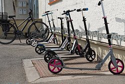 Mehrer Kinderroller und ein Fahrrad stehen entland einer Hauswand in dafür vorgesehenen Halterungen.