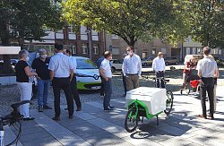 Teilnehmerinnen und Teilnehmer des Werkstattgesprächs zur  umweltfreundlichen Mobilität in der Ortenau stehenmit coronabedingsten Abstand auf einem Platz. Sie tragen fast alle einen Mund-Nasenschutz und unterhalten sich. Zu sehen sind auch geparkte Autos und Fahrräder, Häuser und Bäume entlang einer Straße