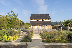 Das Foto zeigt das Streifenhaus in der Römeranlage, die Sonne scheint, blauer Himmel mit einigen Schleierwolken. Im Vordergrund sieht man die schön angelegten Blumenbeete.