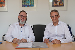 Bürgermeister Schäfer und OB Ibert sitzen an einem Tisch nebeneinander, vor Ihnen liegt eine Unterschriftenmappe. Beide lächeln in die Kamera. 