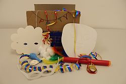Das Foto zeigt die Partybox aus Karton, davor den Inhalt wie zum Beispiel Luftballons, Luftschlangen und Gesichtsmasken aus weißem Papier zum Anmalen.