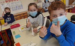 Drei Schüler sitzen mit Maske in der Klasse. Halten ihren Selbsttest in die Höhe und zeigen den hochgestreckten Daumen. 
