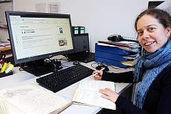 Elise Voerkel sitze an ihrem Schreibtisch vor dem PC mit einer geöffneten Seite der Feuerwehr. Sie lächelt herzlich in die Kamera. 