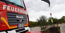 Im Vordergrund ein Feuerwehrfahrzeug von vorne, an dessen Kran eine Atemflasche hängt in gelb, die als Zeitkapsel fungiert. 