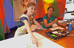 Zwei Mädchen sitzen in einem Zirkuszeelt vor Malutensilien und malen mit Korken und Sprühflasche. 