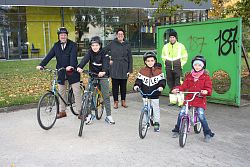 Vier Laufräder besetzt mit einem Mann in schwarzem Mantel, zwei Schülern und einer Schülerin mit rotem Mantel. Im Hintergrund steht eine Frau mit kurzen dunklen Haaren und grauem Mantel, daneben ein Mann in Arbeitskleidung in neongelb/schwarz mit Reflektorstreifen. Alle Personen lächeln und schauen in die Kamera. Im Hintergrund eine Wiese, braunes Laub darauf und eine Glasfassade eines Gebäudes.