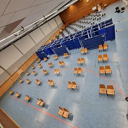 Eine Halle mit blauem Sichtschutz in der Mitte, davor und dahinter stehen paarweise Stühle in großem Abstand zueinander.