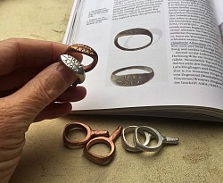 Vor einem aufgeschlagenen Buch mit Abildungen historischer Ringe, hält eine Hand zwei Ringe (bronze und silber). Vor dem Buch liegen vier Rohlinge, zwei in Bronze, zwei in Silber. 