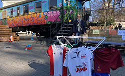 Susanne Schneble steht neben dem Kulturwaggon, davor ein Kleiderständer mit verschiedenen Sporttrikots. 