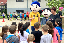 Muck, Minchen, Diana Frei und Kinder der Schutterlindenbergschule stehen zusammmen. Die Kinder hören gespannt zu was Muck und Minchen erzählen.