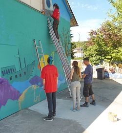 Drei Jugendliche stehen vor einer bunt besprühten Wand, ein weiteres Mädchen steht mit Spraydose auf einer Leiter.